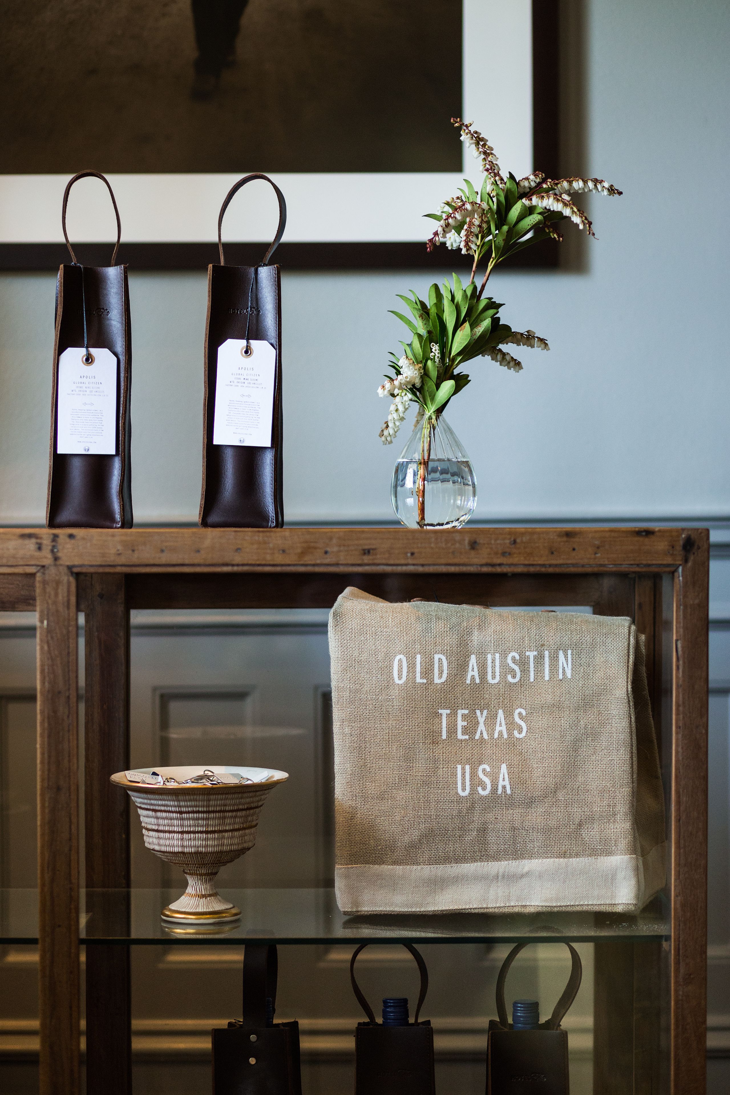 A wooden table with two leather-bound books, a vase with flowers, a bowl, and a burlap bag with "Old Austin Texas USA" text.