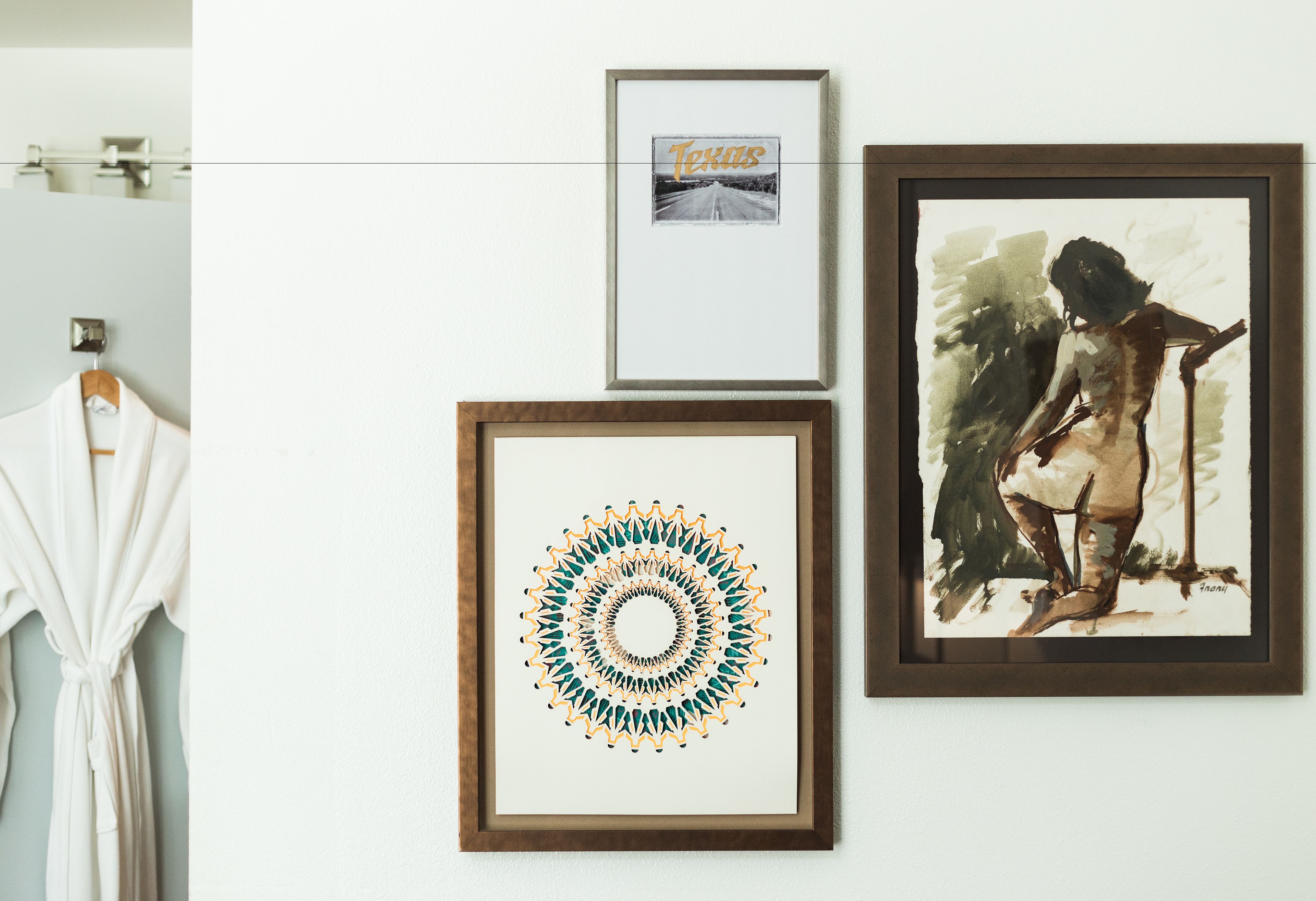 Three framed artworks on a wall, a white robe hanging on a rack, and a mirror.