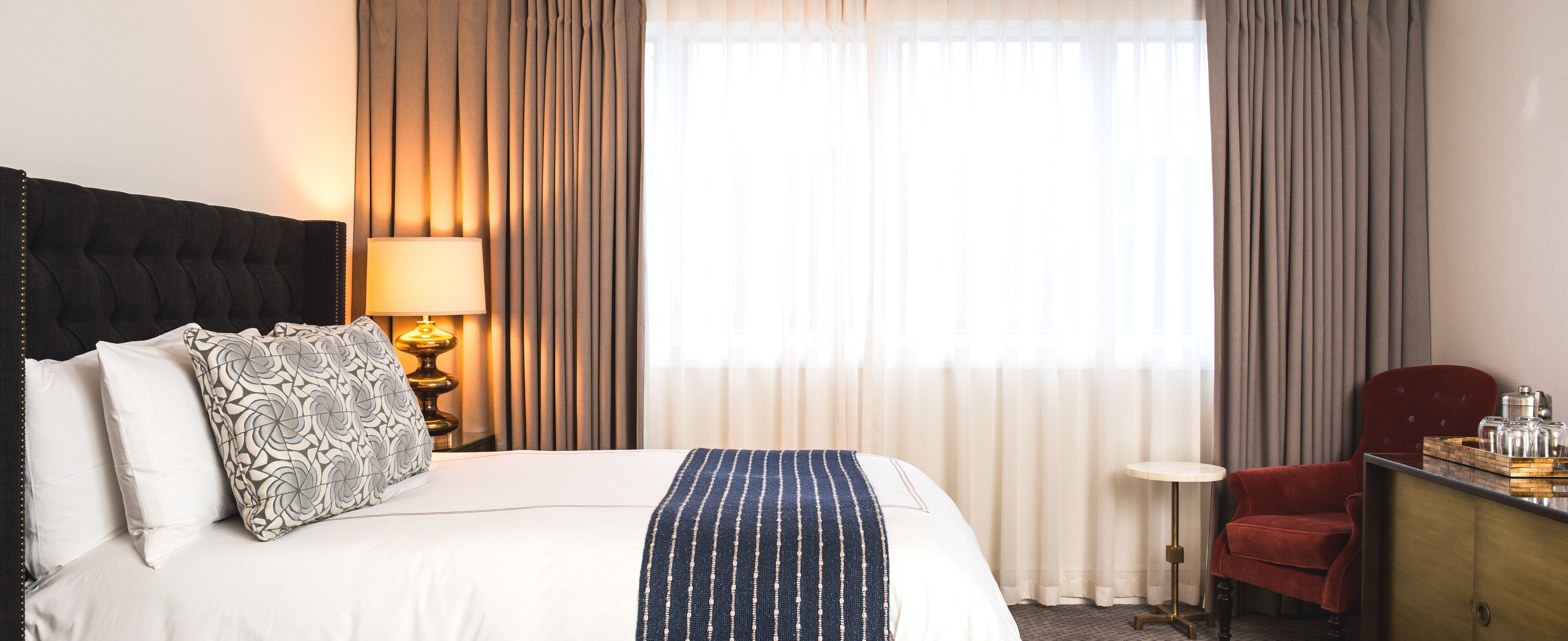 A hotel room with a bed, a bedside lamp, a red chair, and a small table with a tray of items.