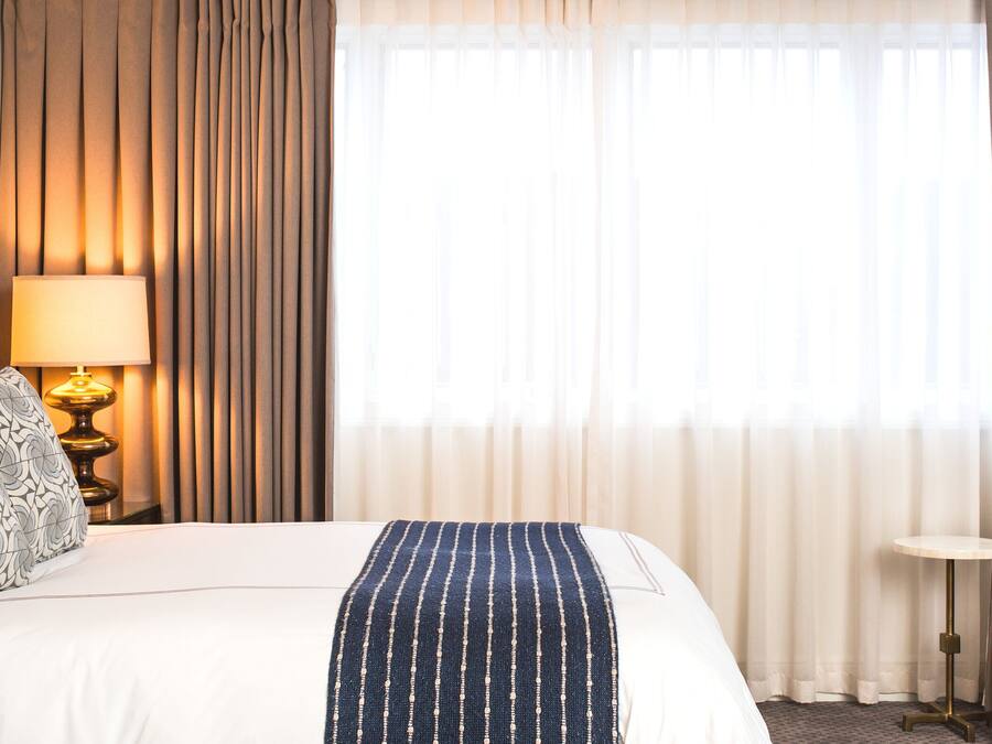Una habitación de hotel con una cama, una lámpara de noche, una silla roja y una mesita con una bandeja de objetos.