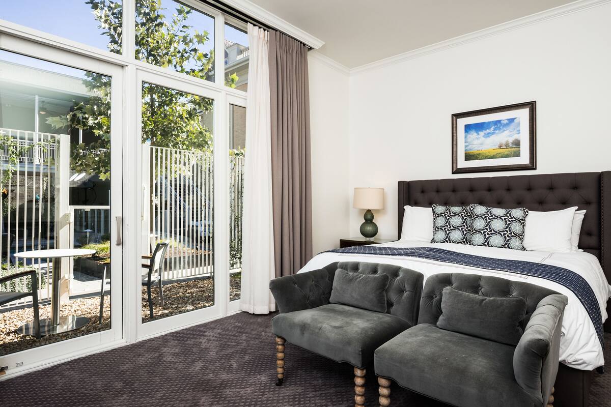 A hotel room with a large window, a bed with a dark headboard, a grey sofa, and a framed picture on the wall.
