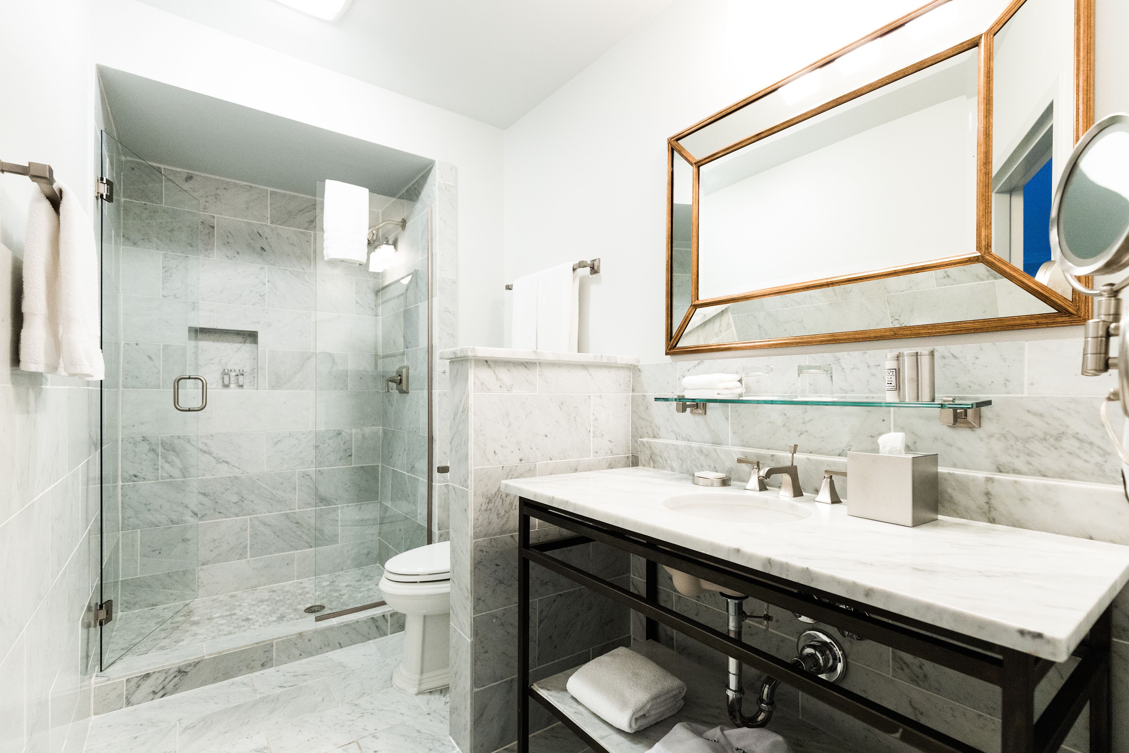 A modern bathroom with a glass-enclosed shower, a white toilet, a marble sink, and a large mirror with a gold frame.