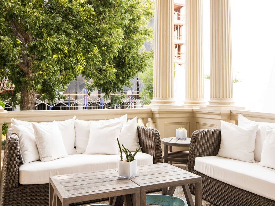 A covered outdoor seating area with a sofa, two tables, and a large window with columns.