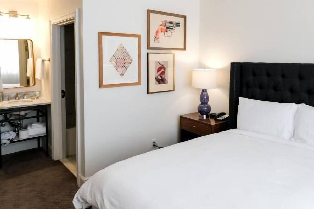 A hotel room with a large bed, a bedside table with a lamp, a small desk with a telephone, a mirror, and two framed pictures on the wall.