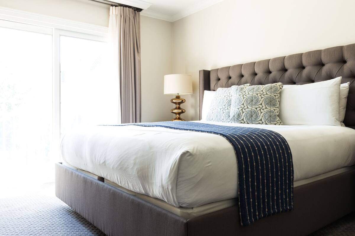 A neatly made bed with a headboard, a bedside lamp, and a window with curtains.