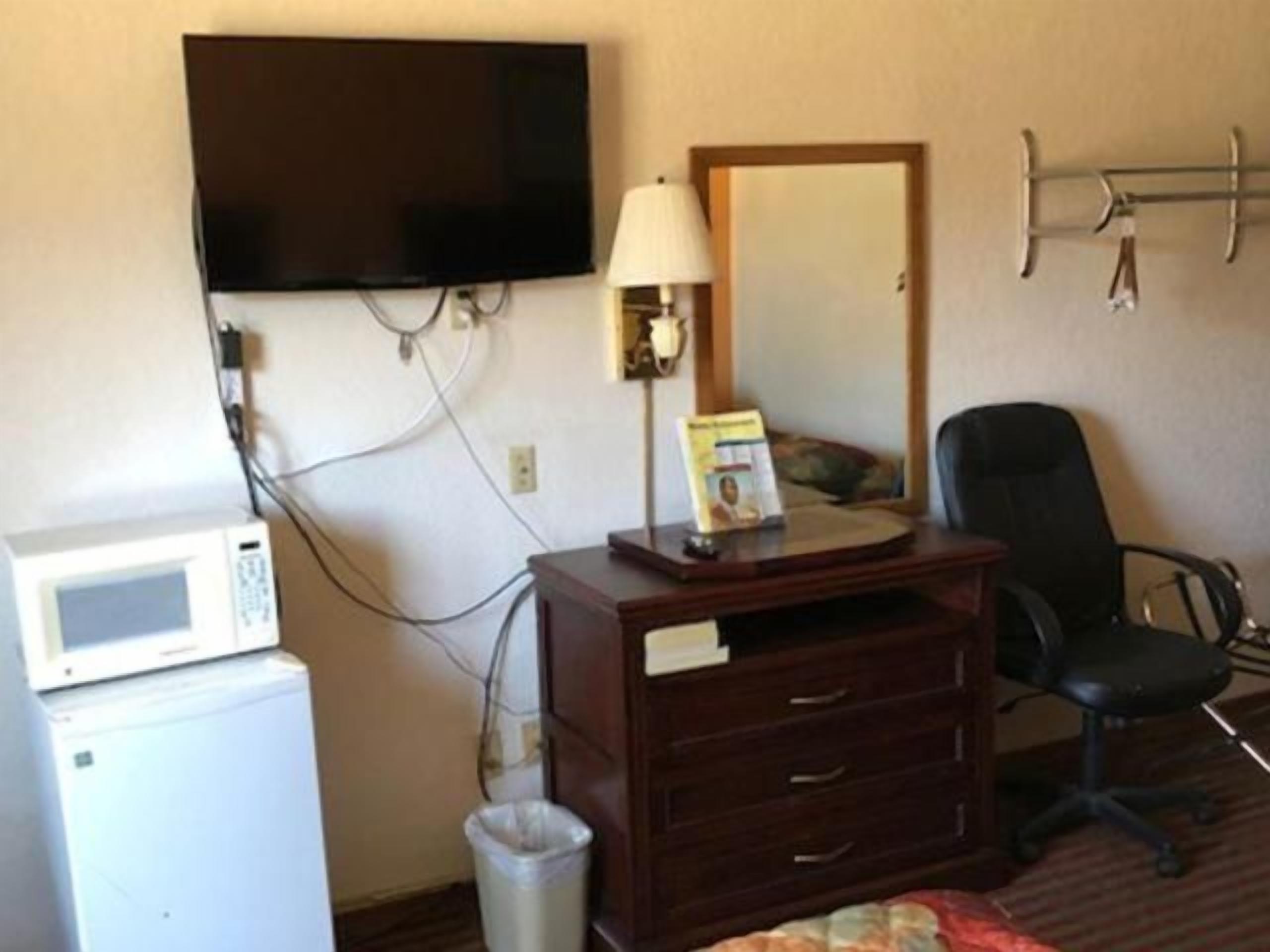 Habitación de hotel con una televisión montada en la pared, un escritorio con una lámpara, una silla, un microondas y un bote de basura.
