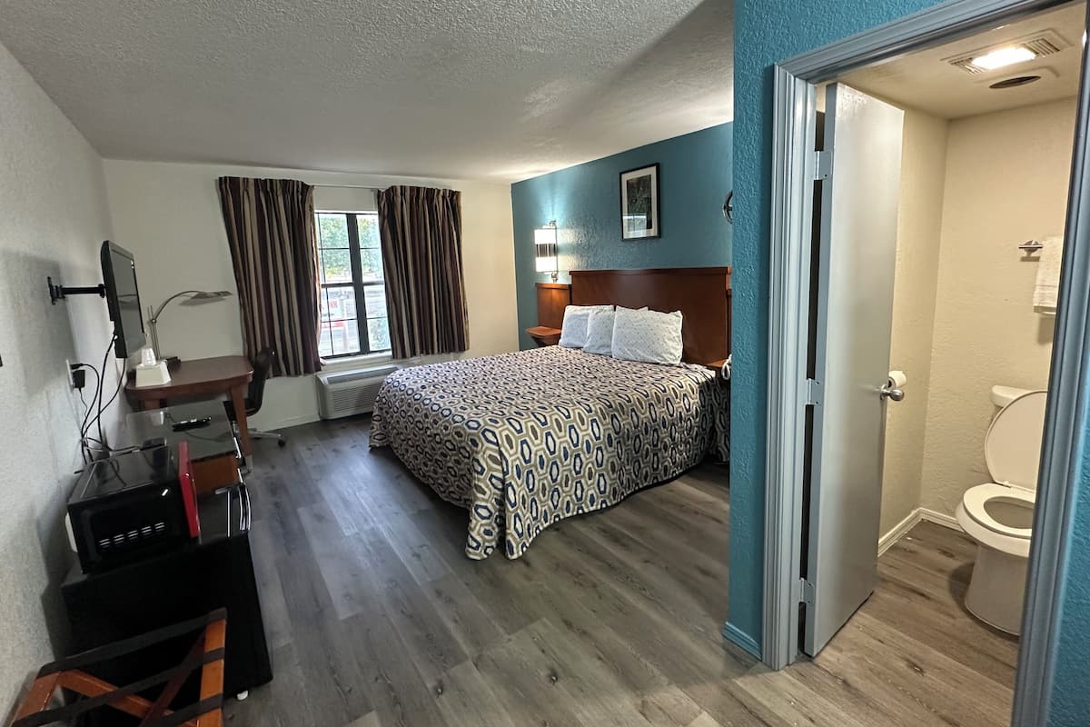 A hotel room with a bed, desk, and computer setup. There is a window with curtains, a bathroom with a toilet, and a door leading to another room.