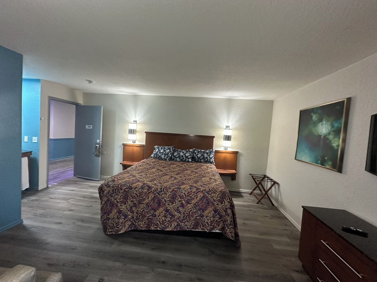 A hotel room with a large bed, two bedside tables with lamps, a dresser, a painting on the wall, and a door leading to another room.