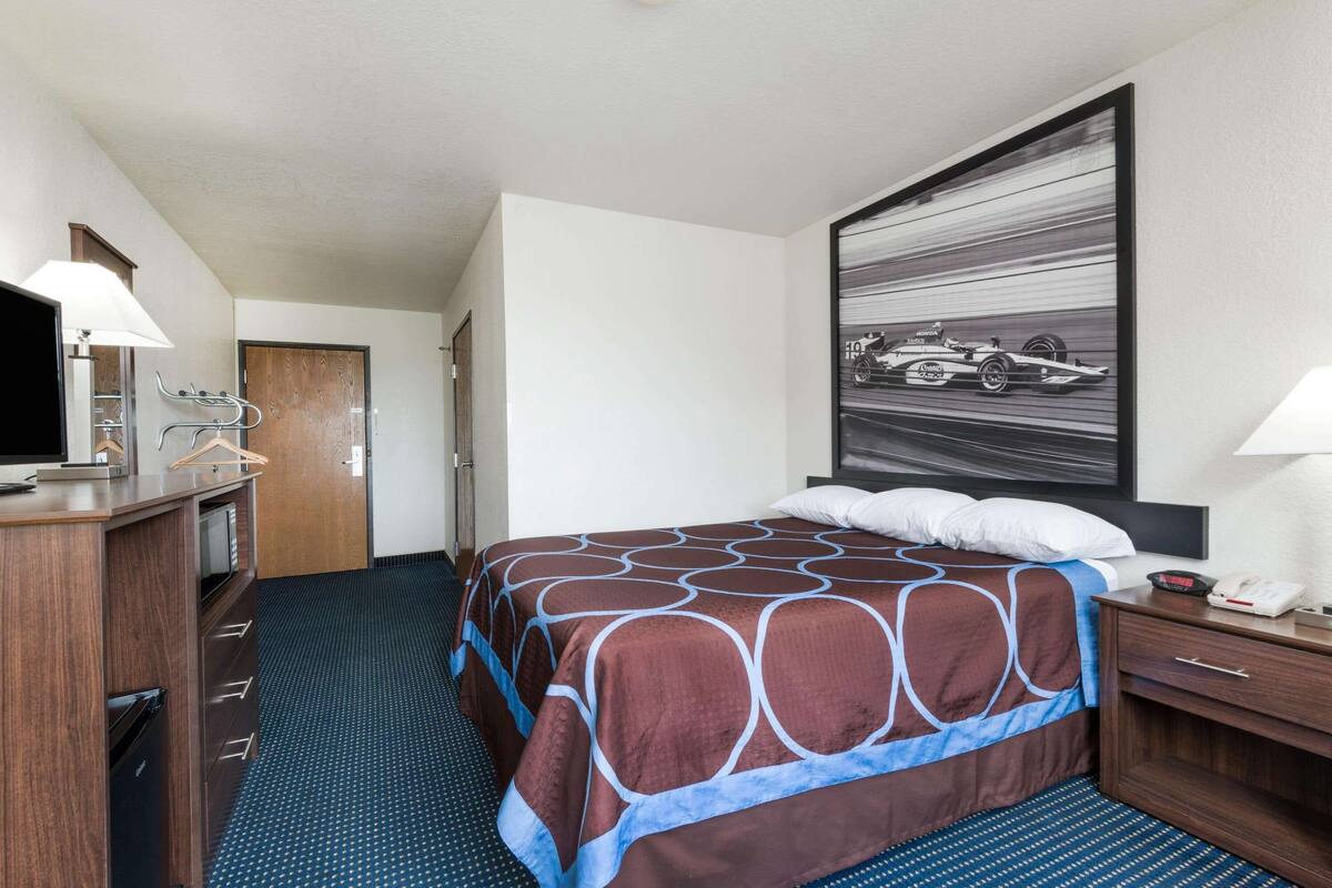 Ein Hotelzimmer mit einem Bett, einem Schreibtisch mit Lampe, einem Fernseher und einem großen Bild in einem Rahmen an der Wand.