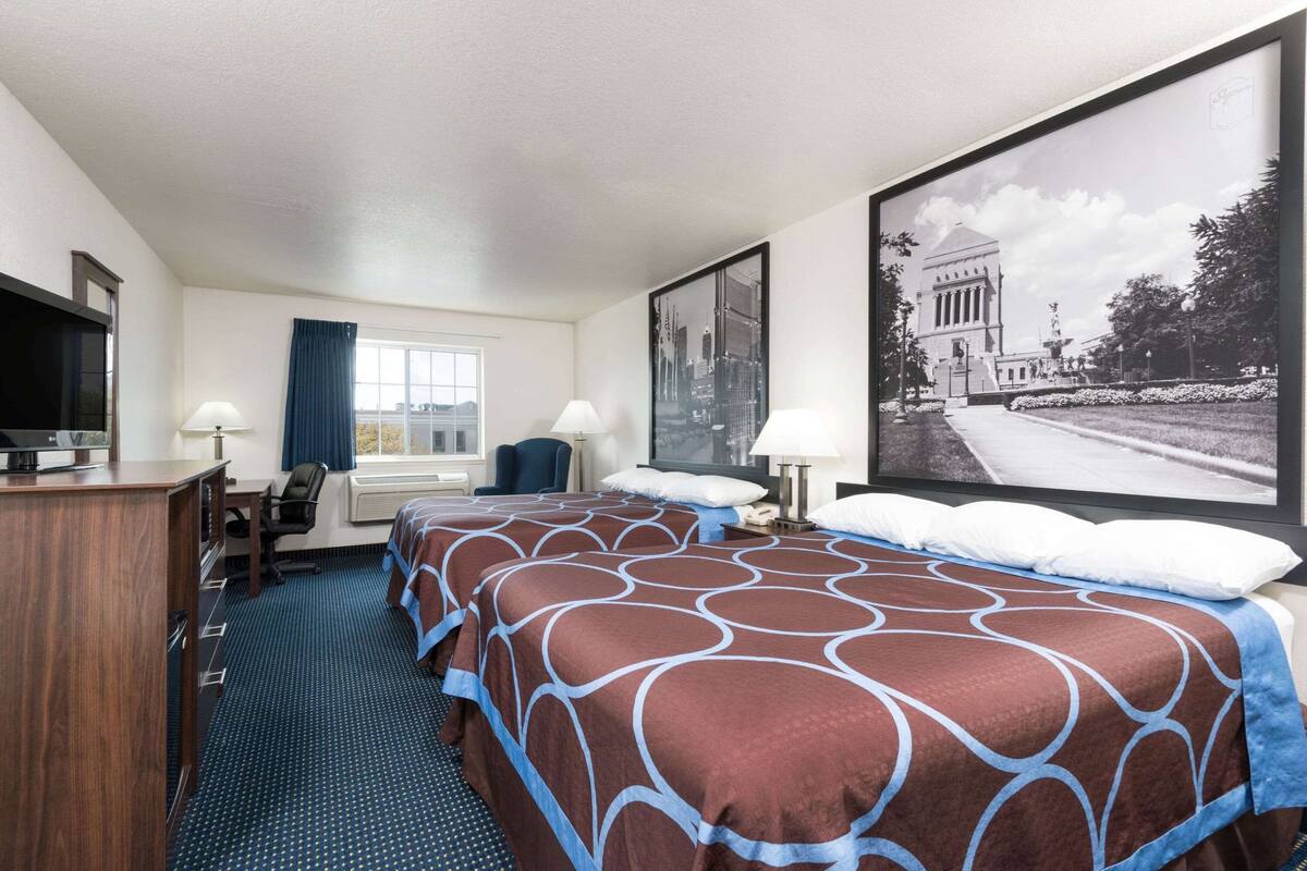 Ein Hotelzimmer mit einem großen Bett, einem Schreibtisch, einem Sessel, einem Fernseher und einem großen gerahmten Bild an der Wand.