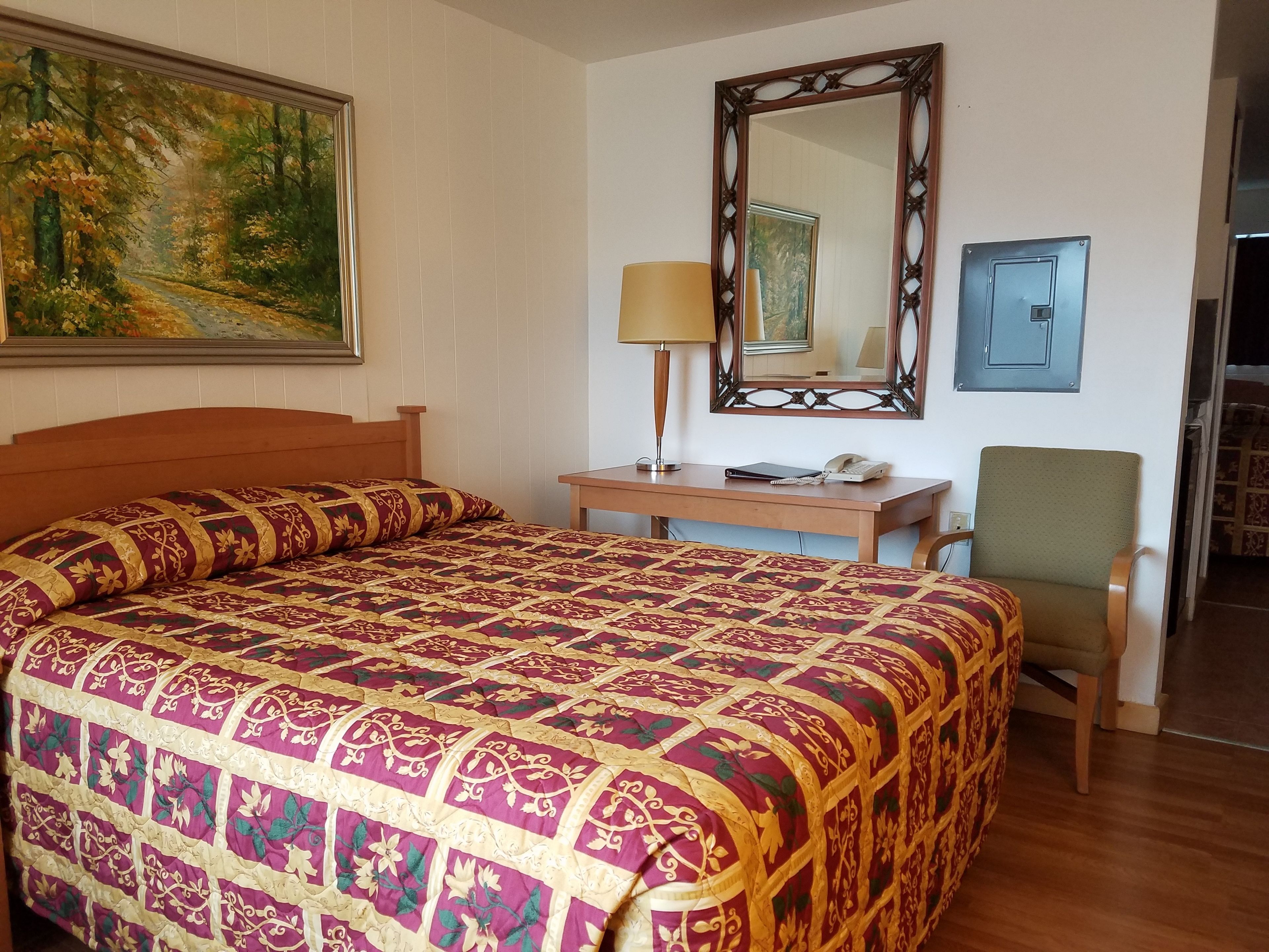 A hotel room with a large bed, a desk, a chair, a lamp, a mirror, and a painting on the wall.