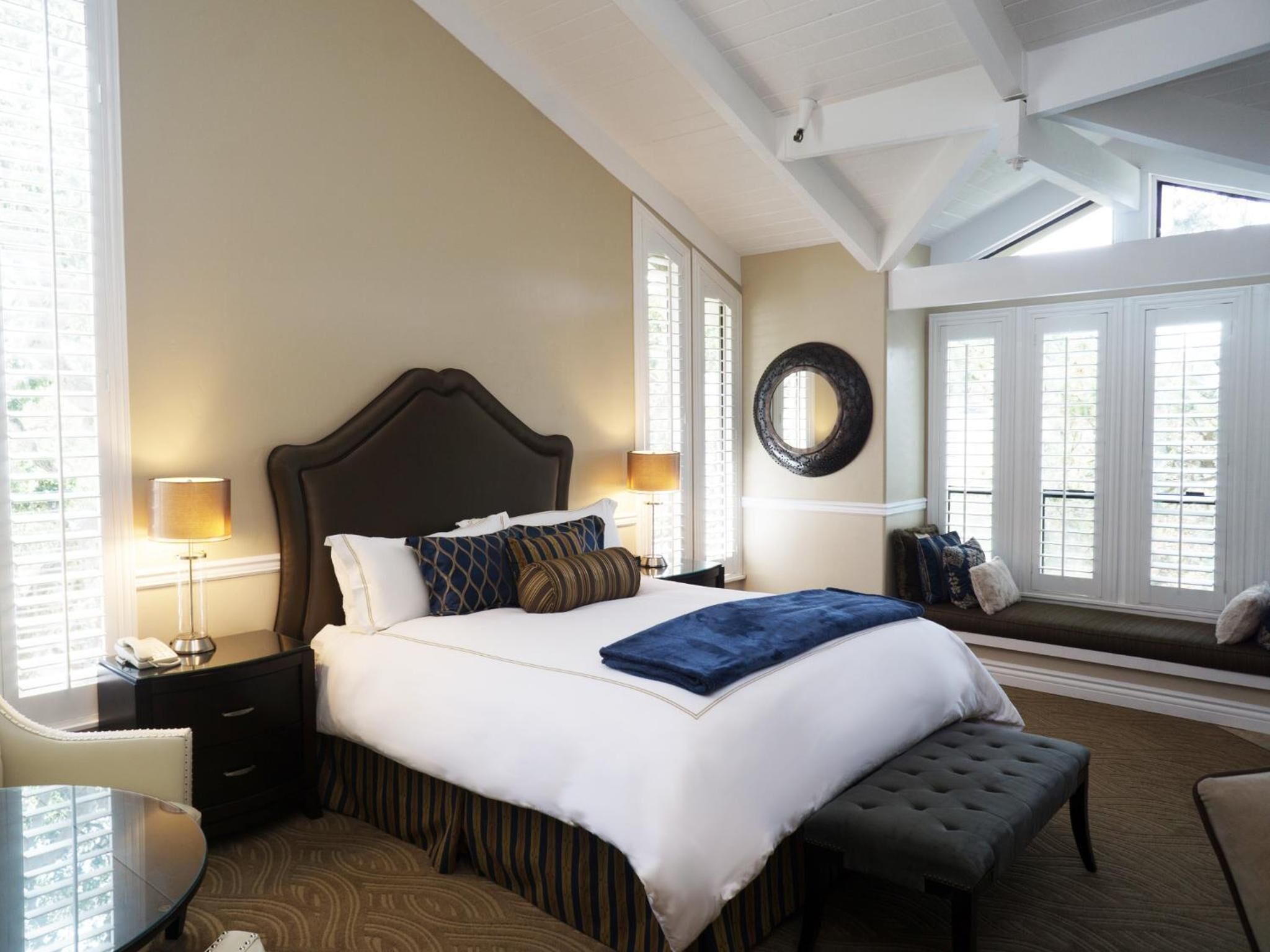 A hotel room with a large bed, a bench, a desk, and a window with blinds.