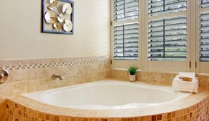 A bathroom with a large bathtub, a wall-mounted artwork, a potted plant, and a window with blinds.