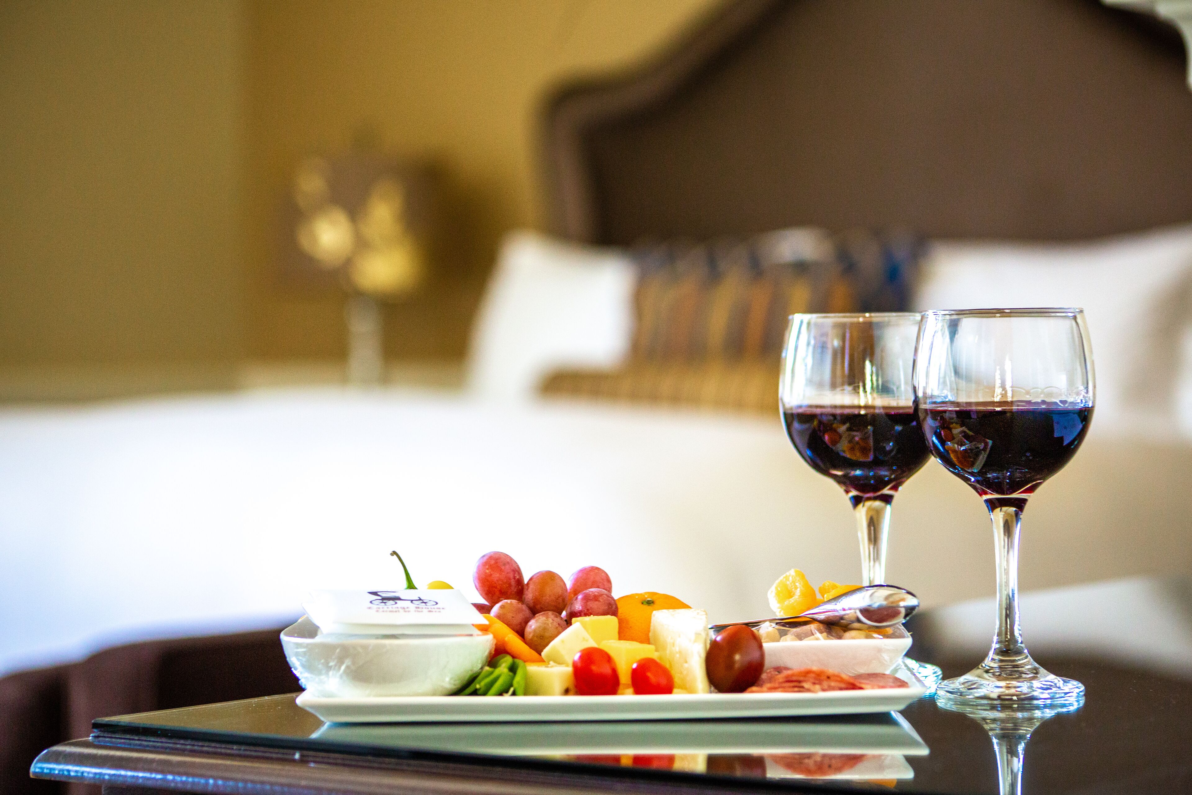 Una bandeja de desayuno ordenada con frutas, queso y un tazón pequeño de yogur, acompañada por dos copas de vino tinto, sobre una mesa en una habitación de hotel.