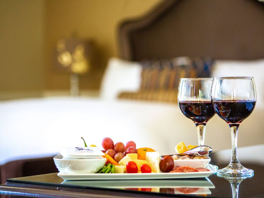 Una bandeja de desayuno ordenada con frutas, queso y un tazón pequeño de yogur, acompañada por dos copas de vino tinto, sobre una mesa en una habitación de hotel.
