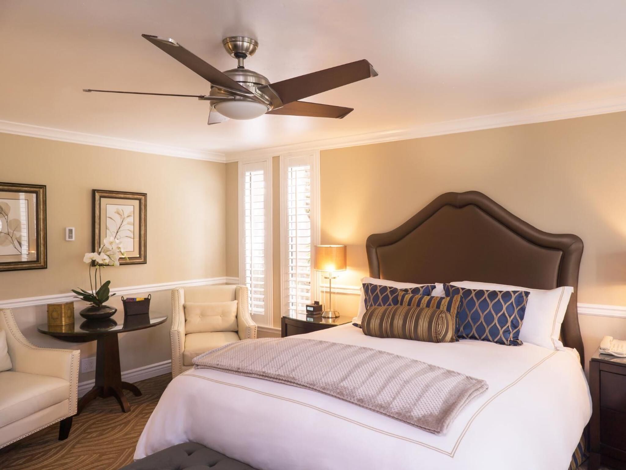 A hotel room with a large bed, a ceiling fan, a small table with a lamp, a chair, and two framed pictures on the wall.