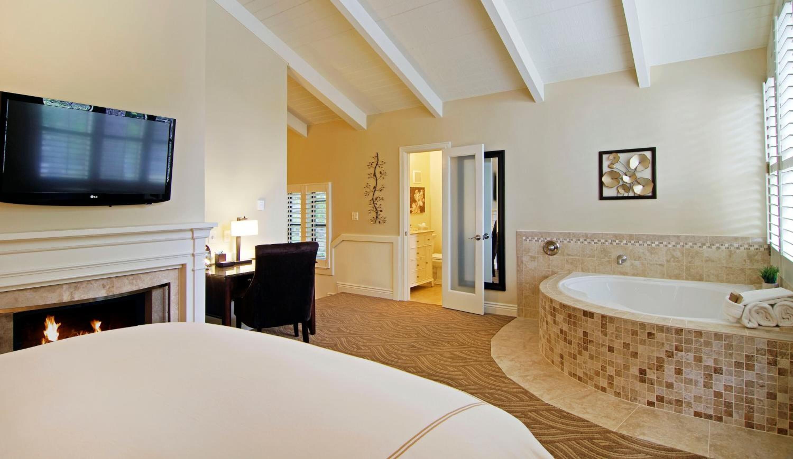 A hotel room with a fireplace, a large bed, a sitting area with a chair, a television, a bathroom with a bathtub, and a door leading to another room.