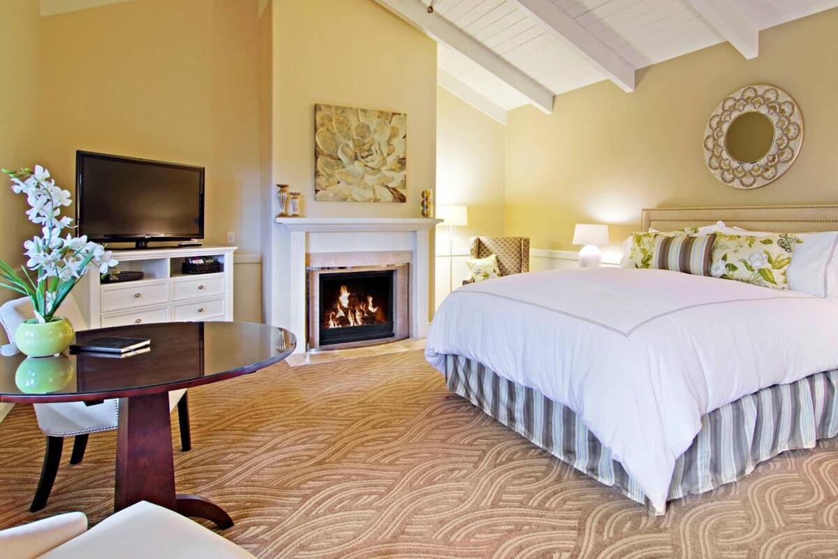 Ein Hotelzimmer mit Bett, Fernseher, Kamin, Tisch mit Blumenvase und einem gemusterten Teppich.