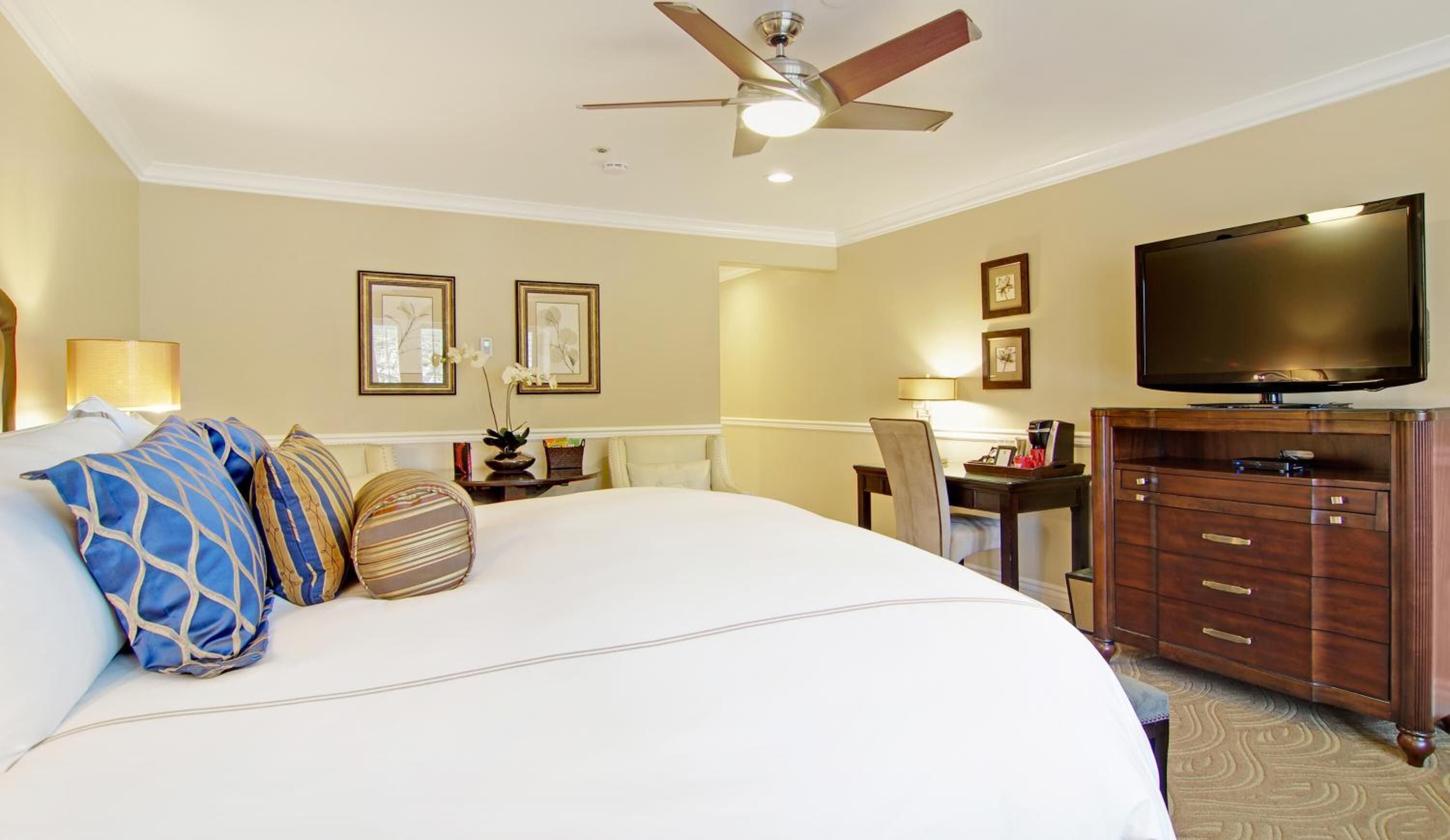 A hotel room with a large bed, a desk with a chair, a television, and a ceiling fan.