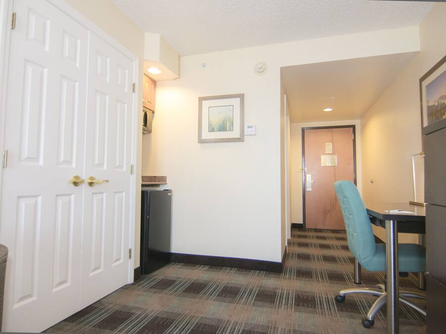 A hotel hallway with a carpeted floor, a white door, a framed picture on the wall, and a desk with a chair.