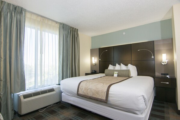 A hotel room with a large bed, a bedside table, a nightstand, a wall-mounted lamp, and a window with curtains.