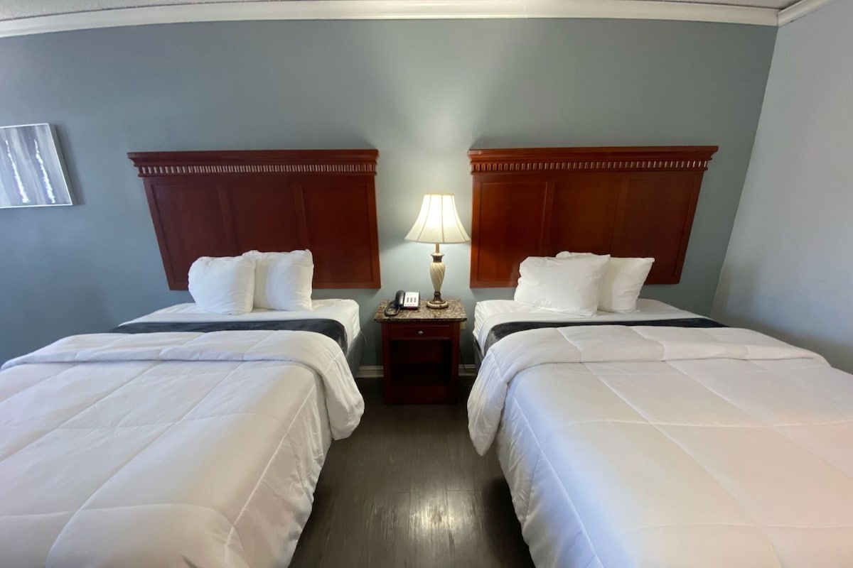 A hotel room with two beds, a nightstand with a lamp, and a wall-mounted phone.