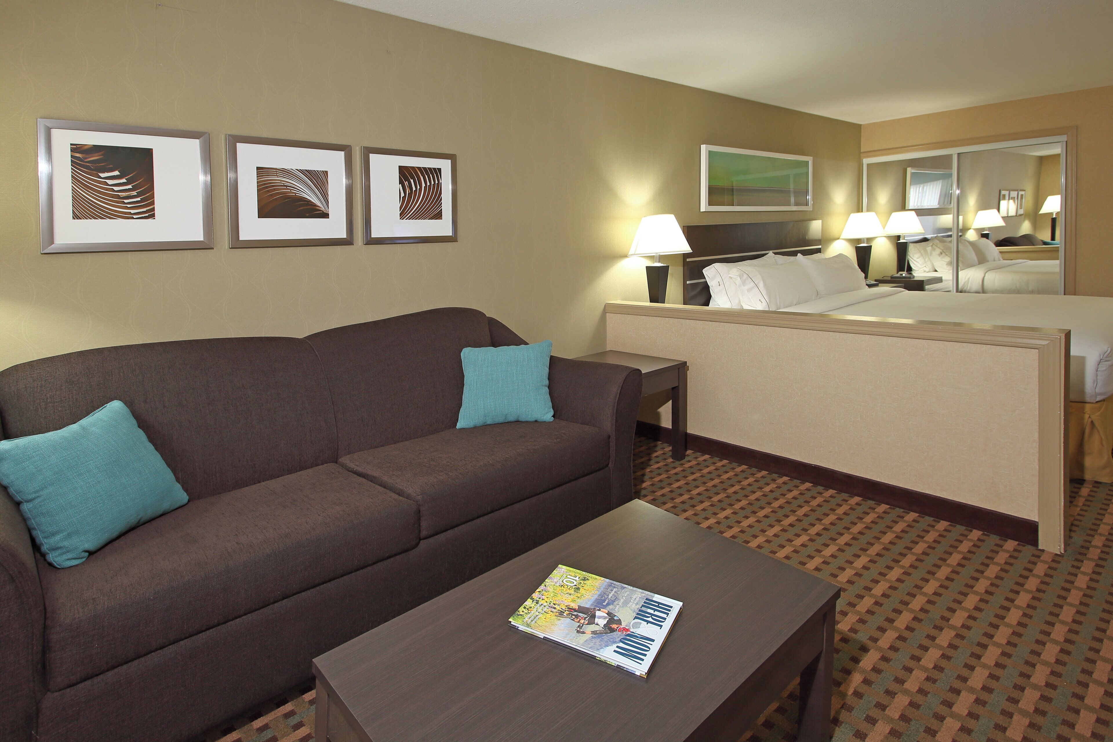 Una habitación de hotel con un sofá marrón, una mesita con una revista, una cama con ropa blanca y tres cuadros en la pared.