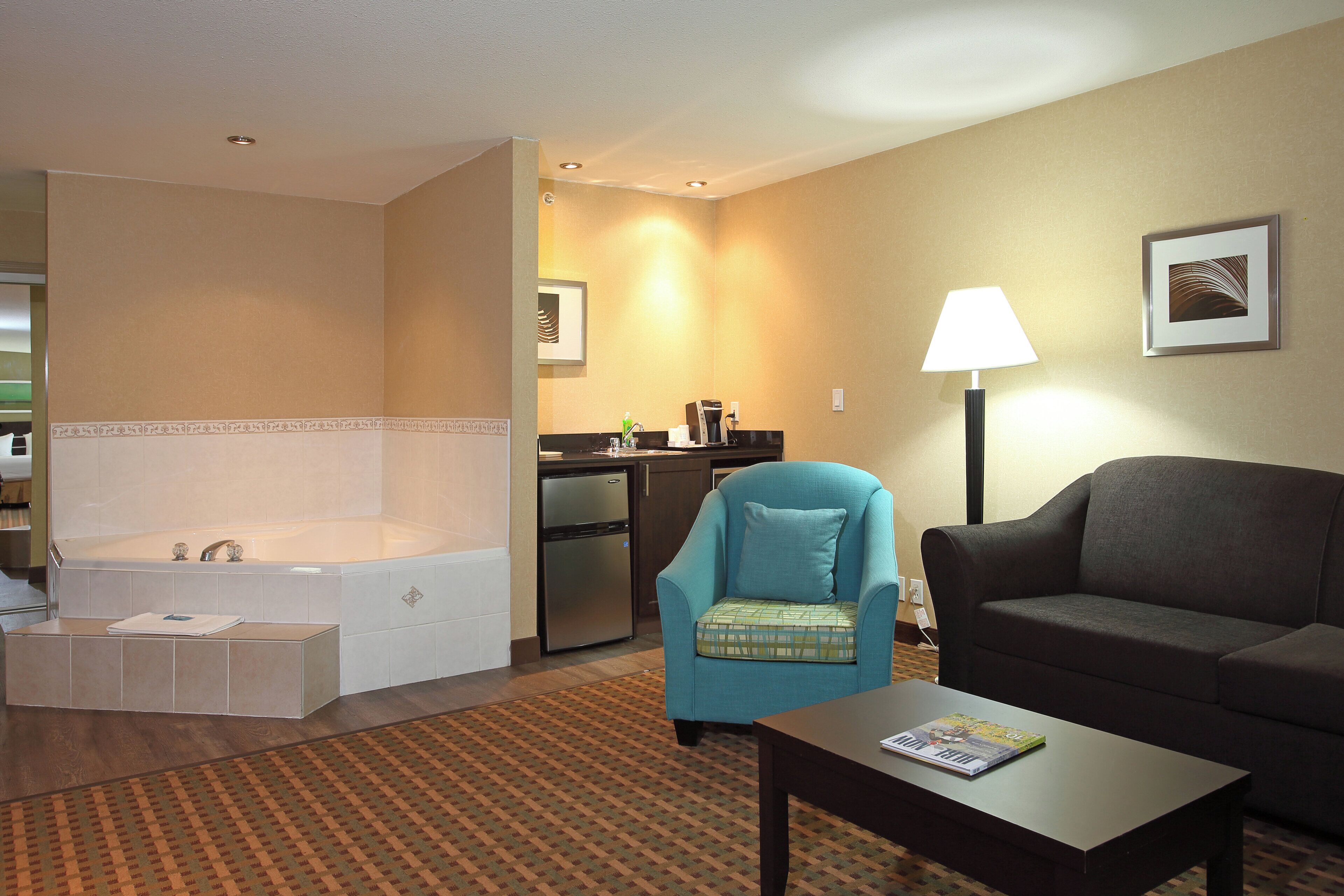 Ein Hotelzimmer mit Wohnbereich, einer Couch, einem Couchtisch, einem Sessel, einer Lampe, einem Bilderrahmen, einer Küchenzeile und einer Badewanne.