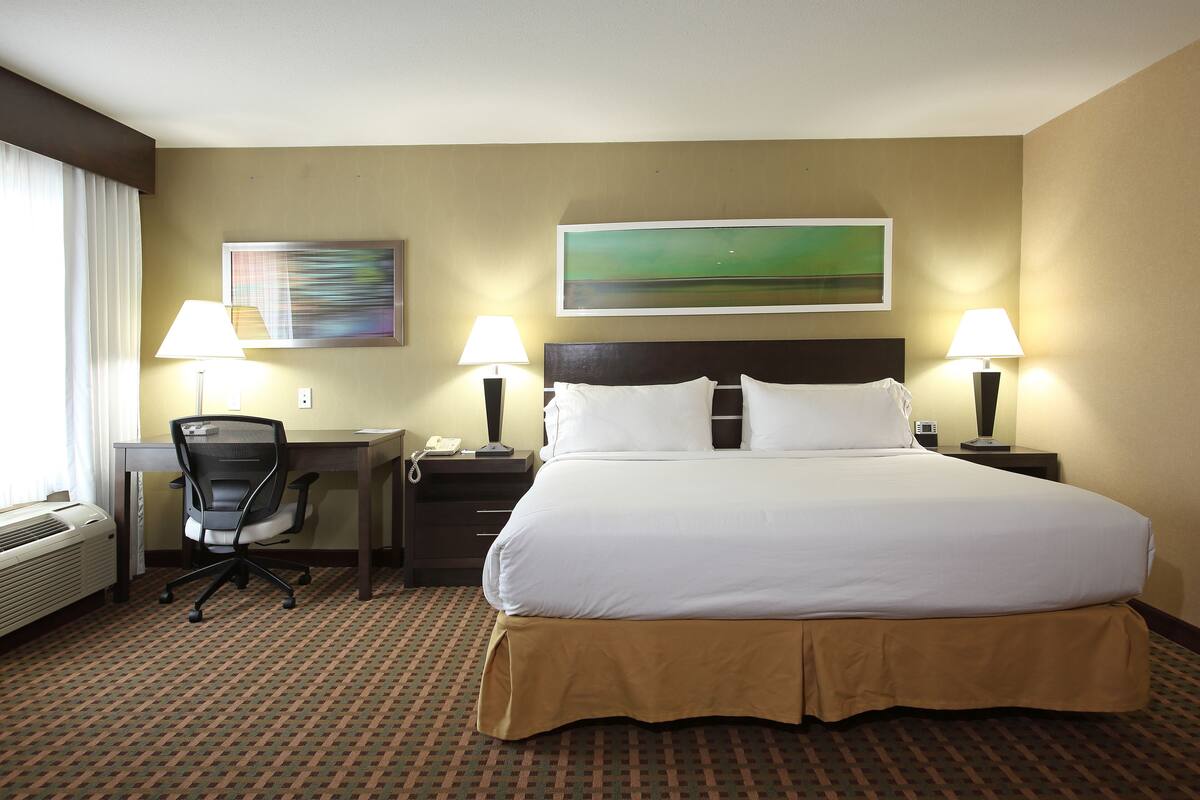 A hotel room with a large bed, two bedside tables with lamps, a desk with a chair, a telephone, and a painting on the wall.