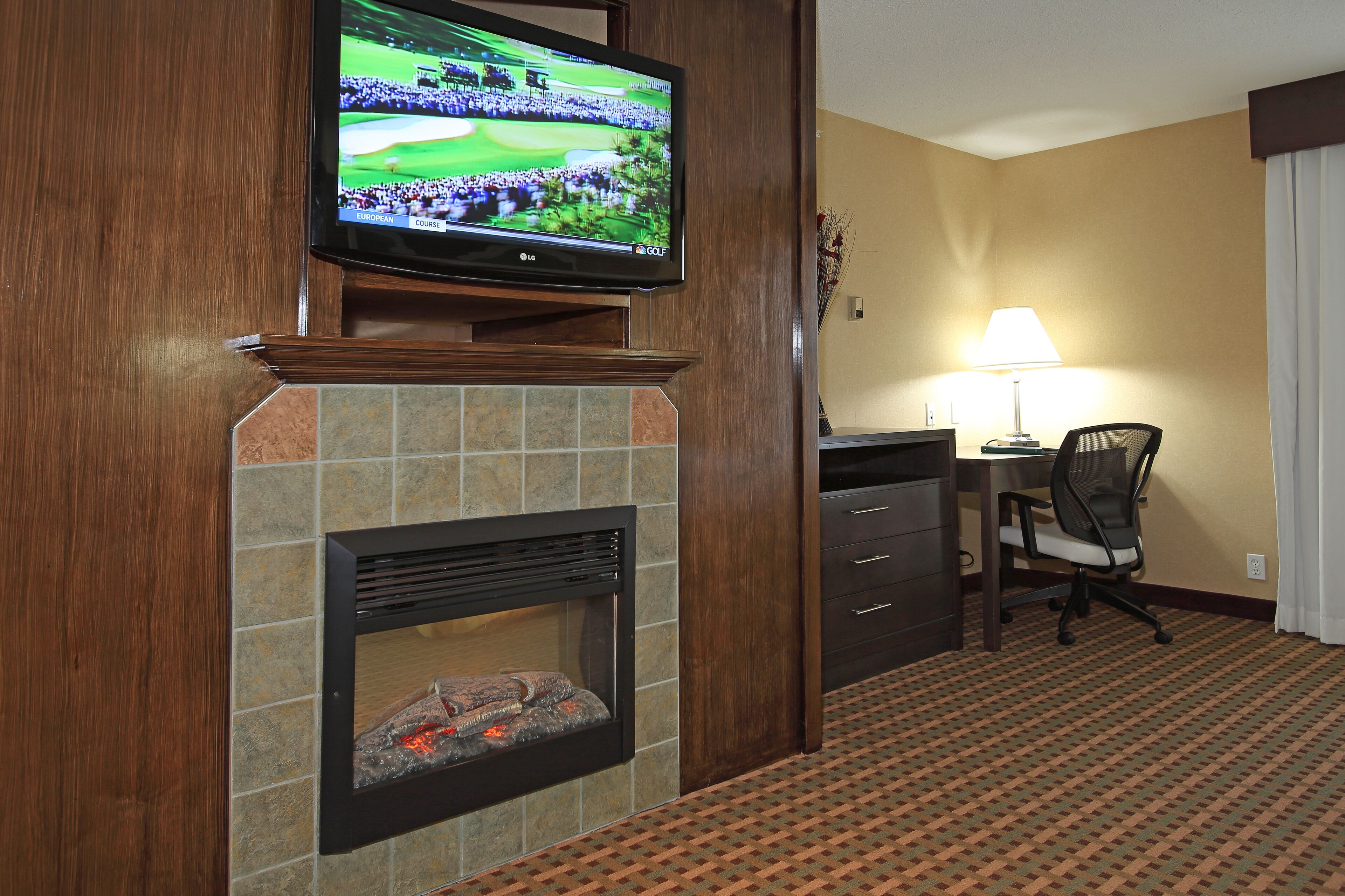 A hotel room with a television mounted above a fireplace, a desk with a chair, and a lamp.