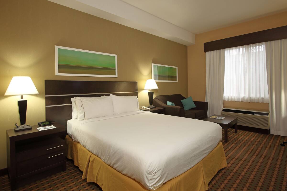 A hotel room with a large bed, two bedside tables with lamps, a chair, a window with curtains, and two framed pictures on the wall.