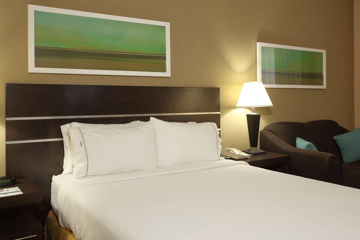A hotel room with a large bed, a nightstand with a lamp, a chair, and two framed pictures on the wall.