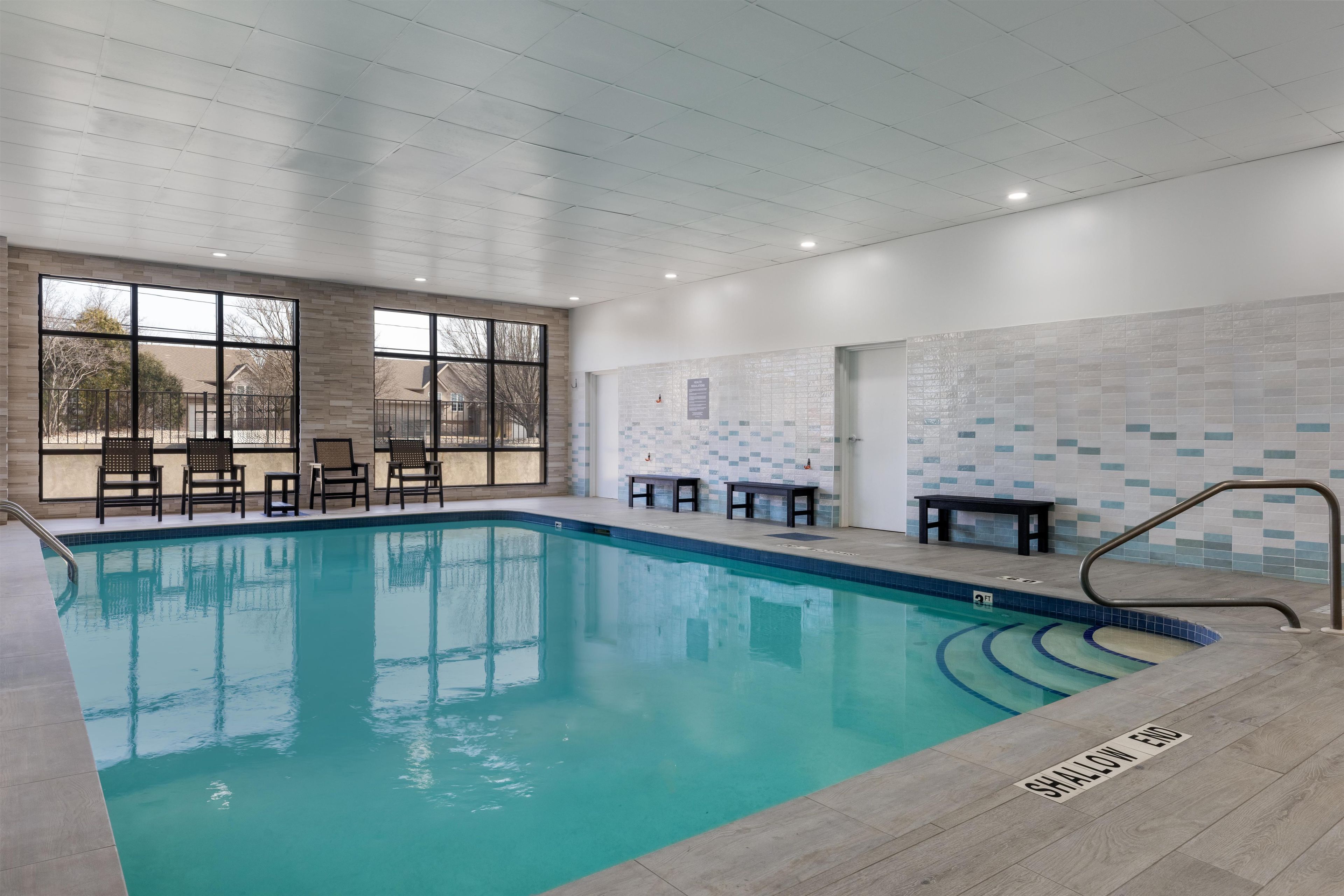 An indoor swimming pool with clear water, surrounded by lounge chairs and tables, and a view of the outside through large windows.