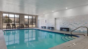 An indoor swimming pool with clear water, surrounded by lounge chairs and tables, and a view of the outside through large windows.