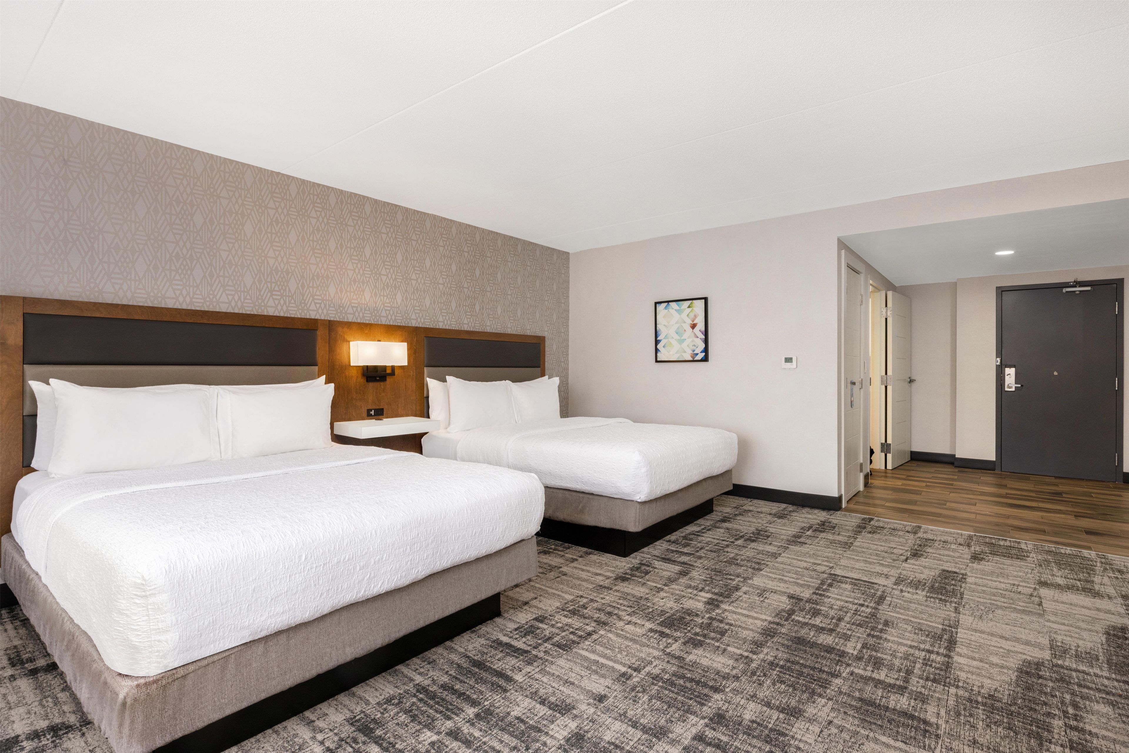 Habitación de hotel con dos camas, cabecero, mesita de noche, lámpara, una obra de arte colgada en la pared y suelo alfombrado.