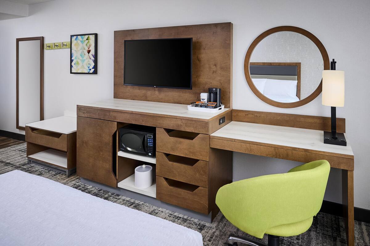 A hotel room with a bed, a desk with a flat-screen TV, a microwave, a coffee maker, a lamp, and a green chair.