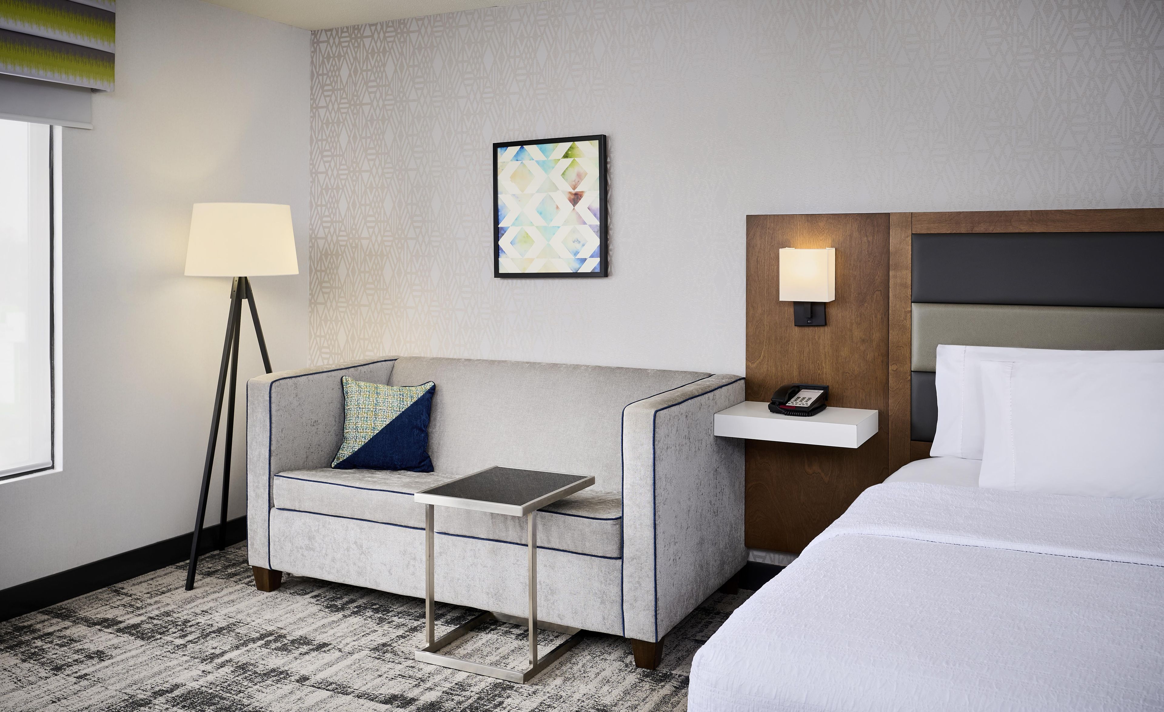 A hotel room with a sofa, a bedside table, a lamp, a phone, and a bed.