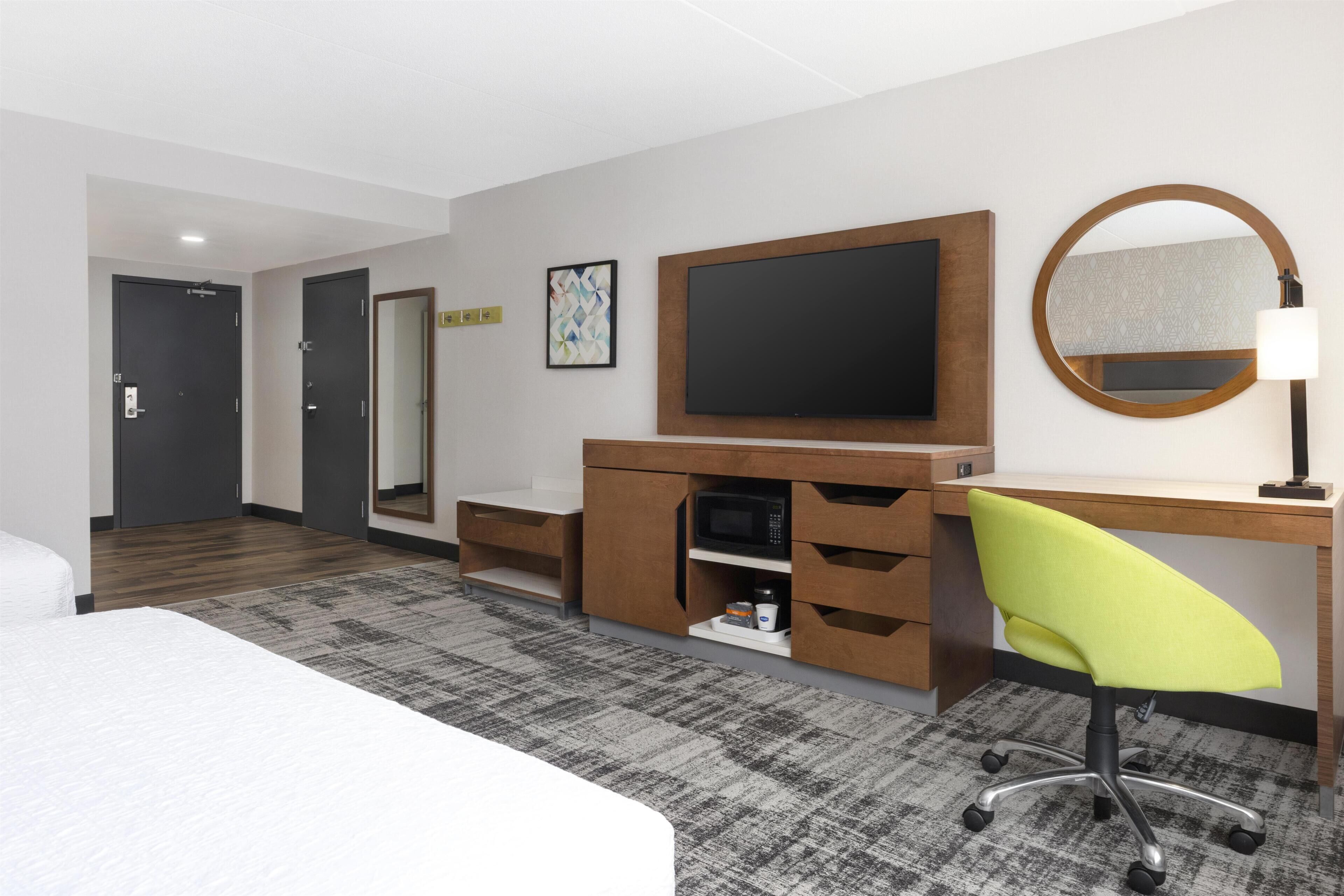 Habitación de hotel con cama, escritorio, silla, televisor, microondas y espejo.