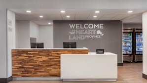 A reception desk with a 'Welcome to the Heartland Province' sign, a wooden reception desk, a white reception desk, and a minibar refrigerator. - Hampton Inn & Suites by Hilton Windsor (Windsor)