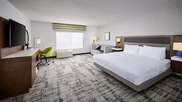 Een hotelkamer met een groot bed, een bureau met een flatscreen televisie, een stoel, een nachtkastje met een lamp en een raam met zonwering.