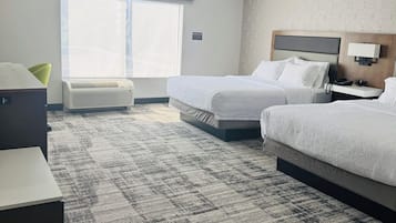 A hotel room with two beds, a desk, a chair, a window with blinds, an air conditioning unit, and a patterned carpet.