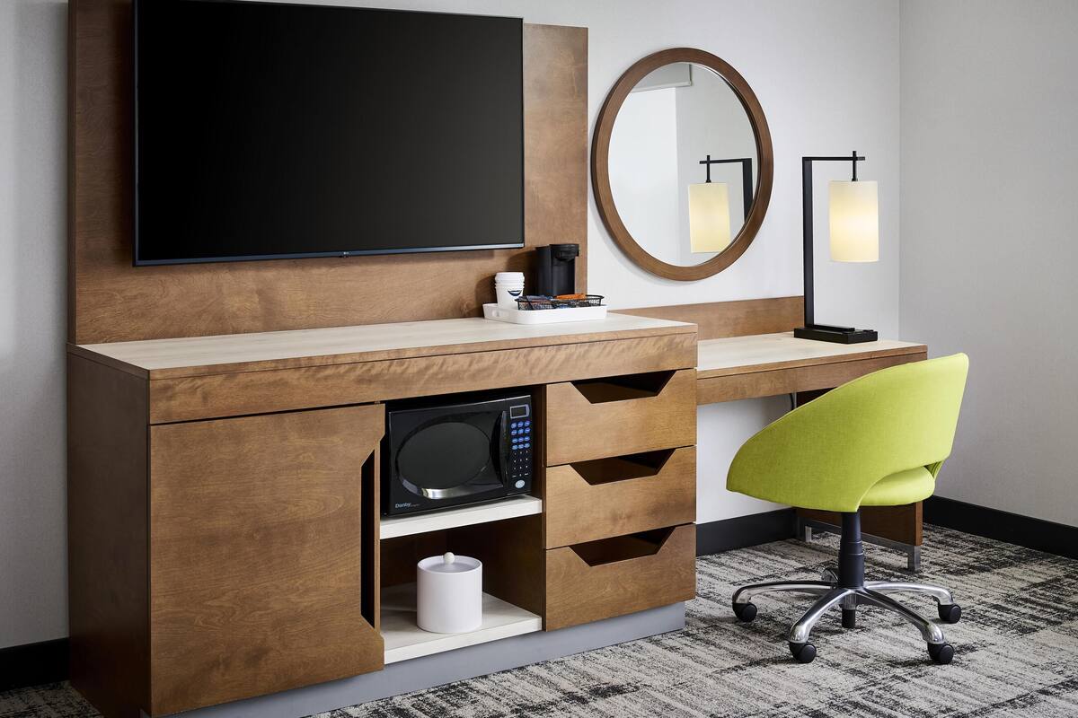 A modern hotel room with a flat-screen TV, a round mirror, a desk with a lamp, a microwave, and a bright green chair.