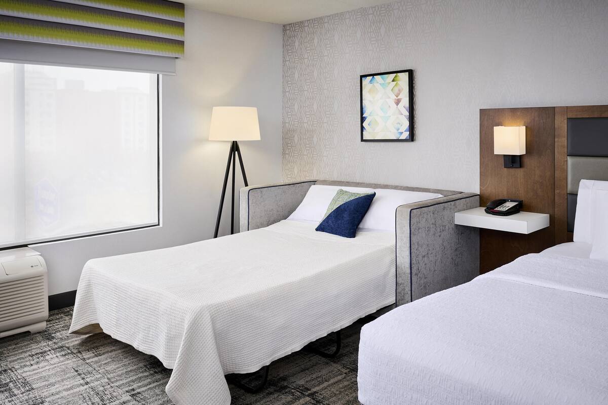 A hotel room with a single bed, a bedside table with a phone, a floor lamp, and a window with blinds.