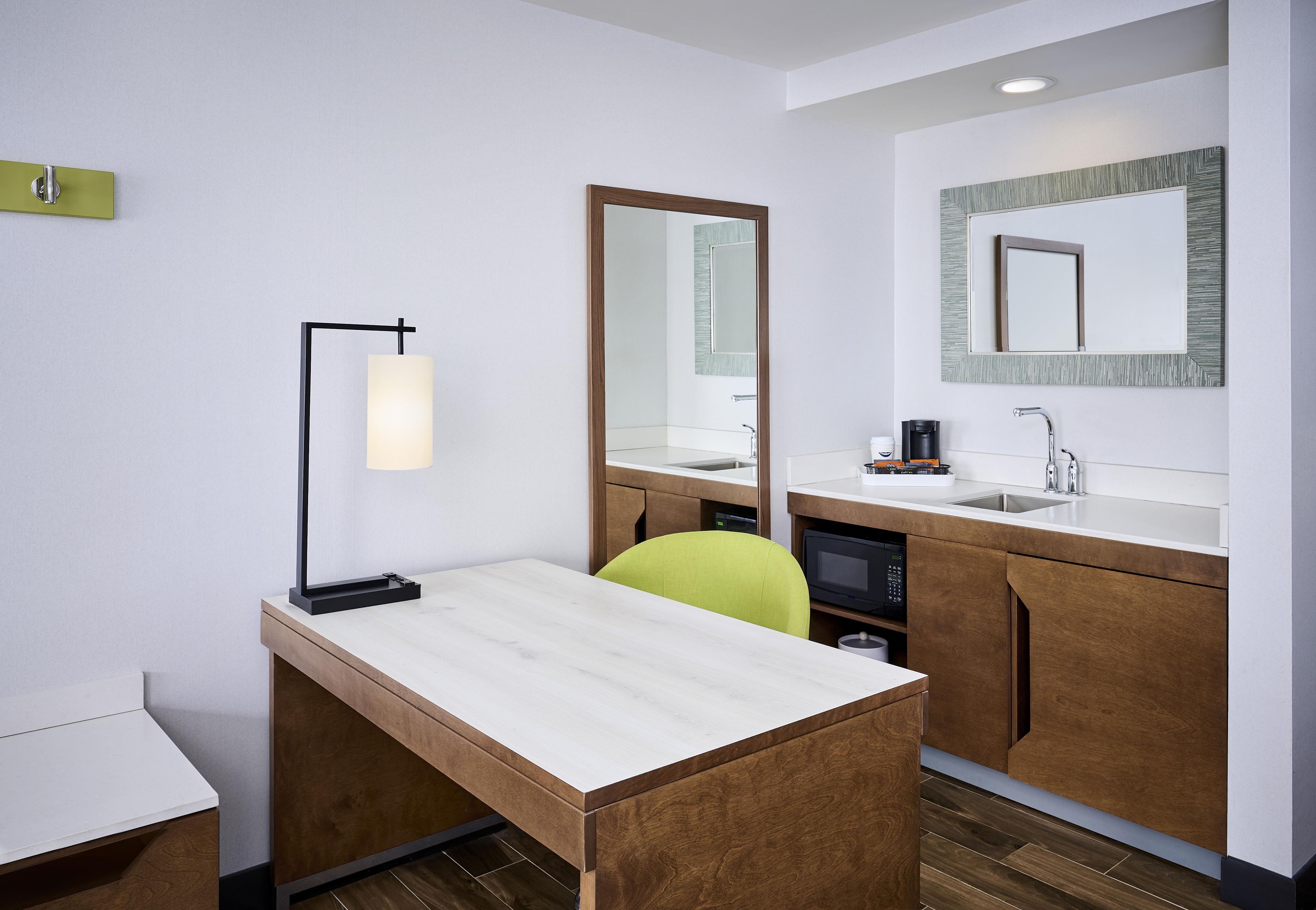 Habitación de hotel con un escritorio de madera, una silla, lámpara de escritorio, espejo, lavamanos y microondas.