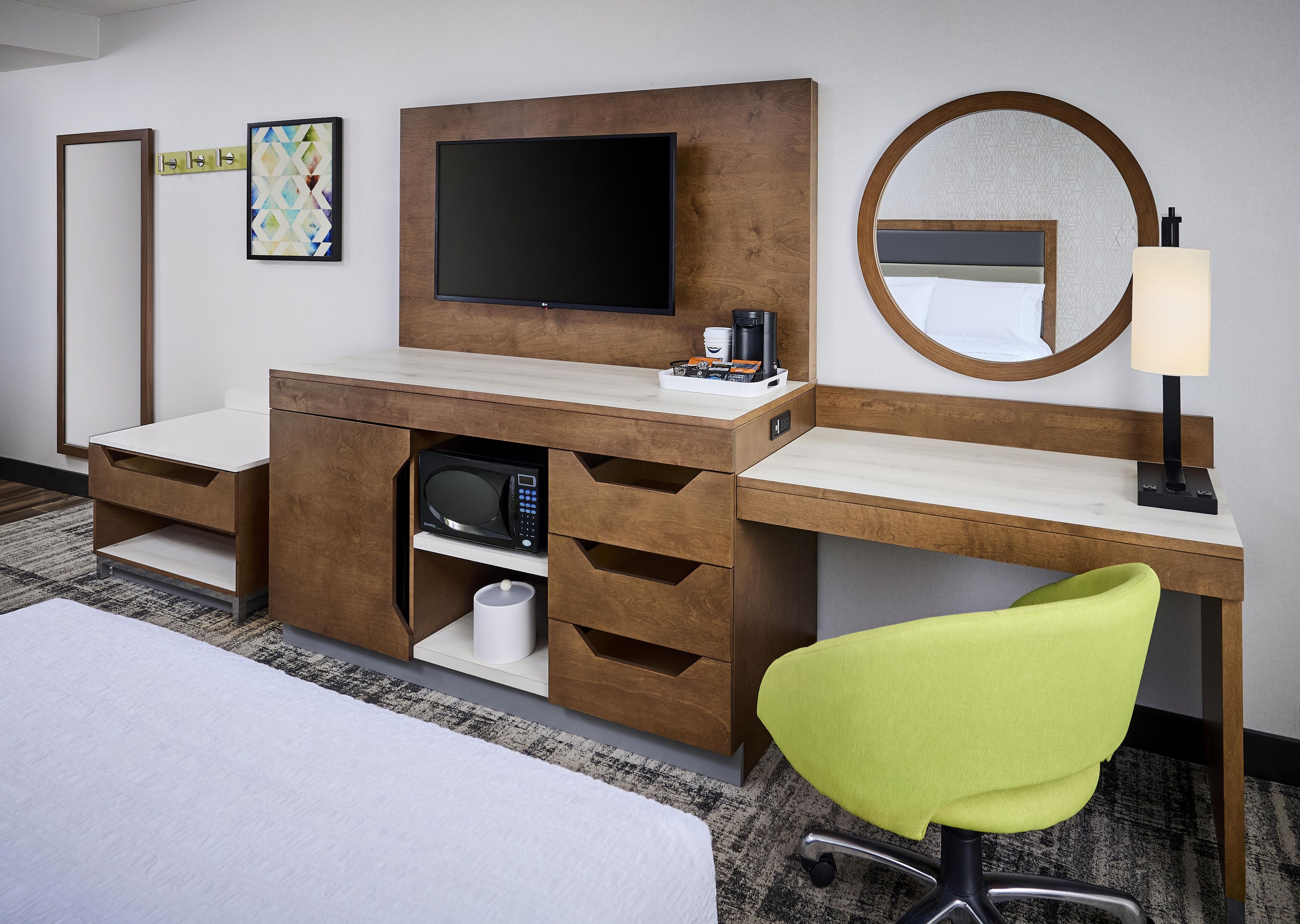 A hotel room with a bed, a desk with a flat-screen TV, a microwave, a coffee maker, a lamp, and a mirror.