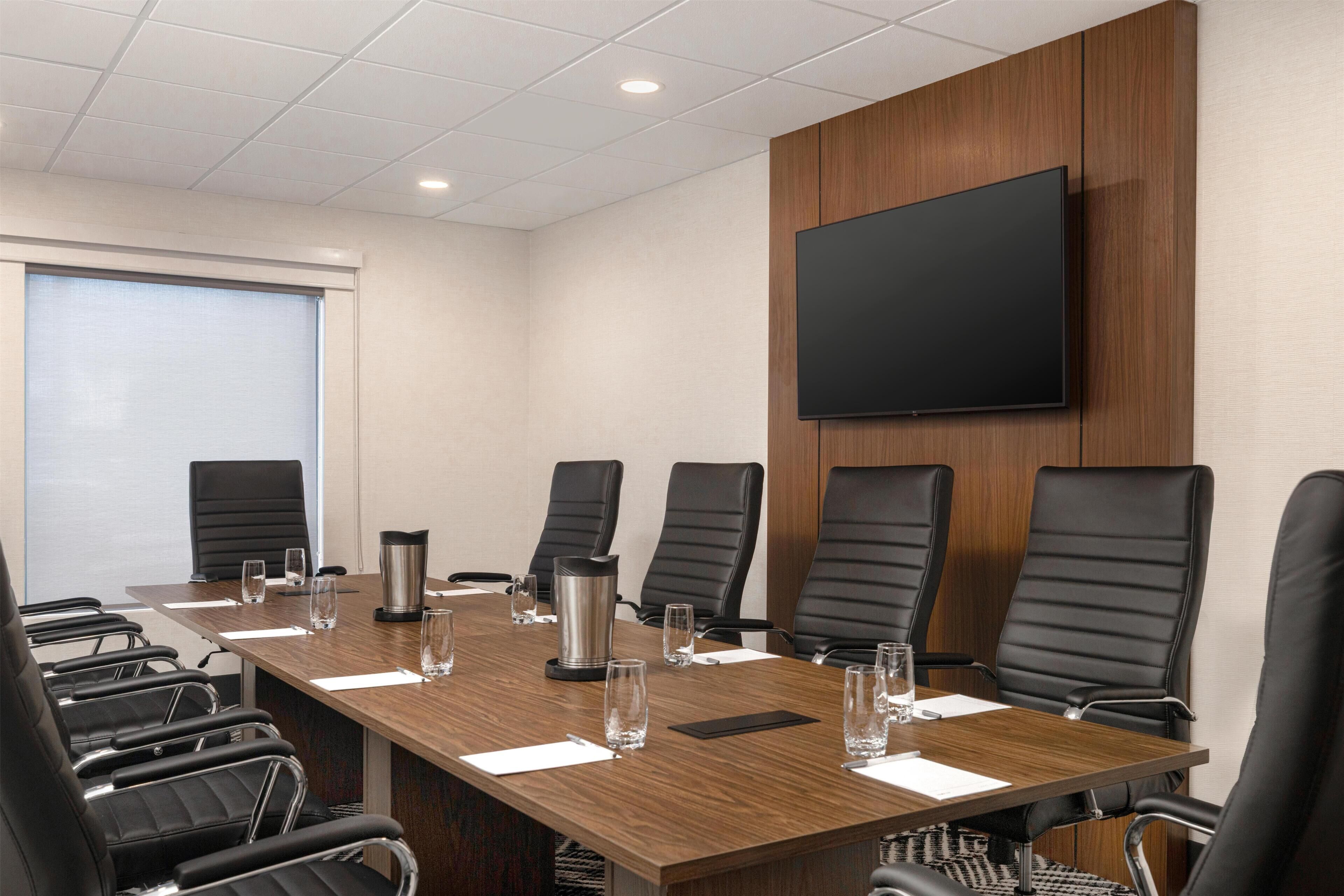 A conference room with a long table, black chairs, a flat-screen TV, and a window with blinds.