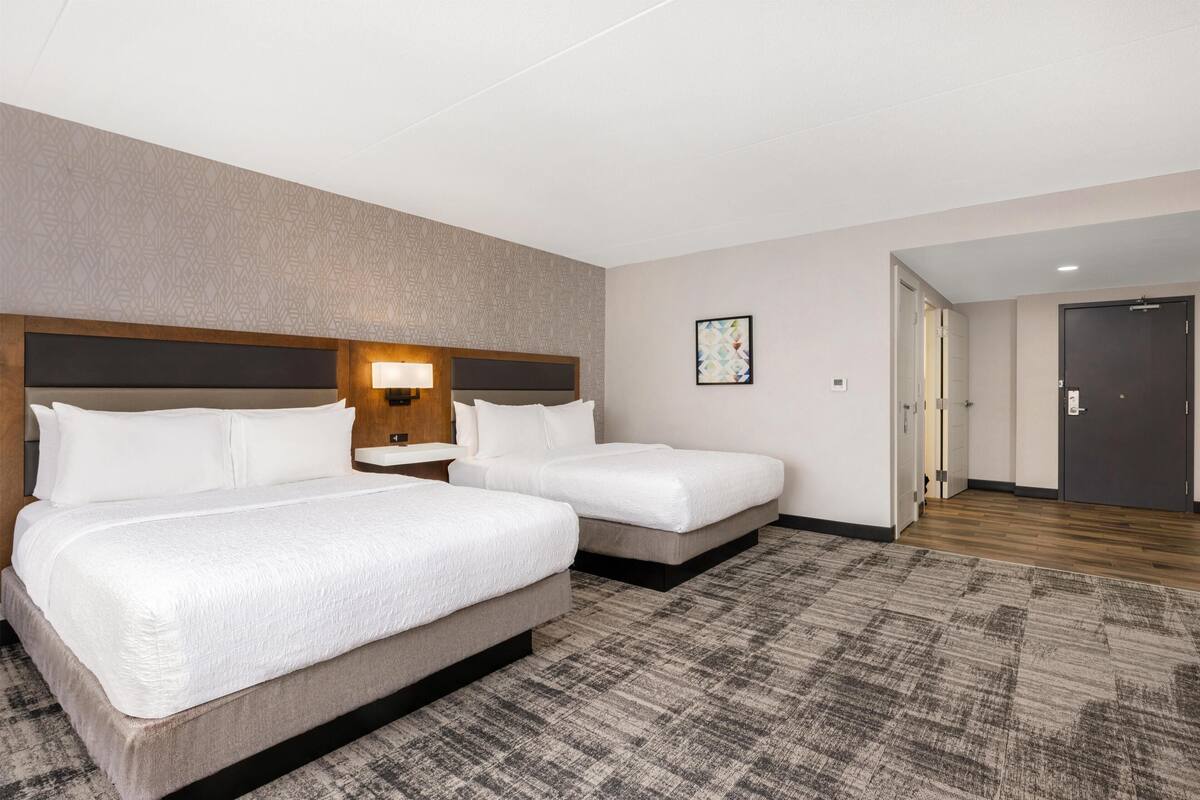 A hotel room with two beds, a headboard, a bedside table, a lamp, a framed picture on the wall, and a carpeted floor.