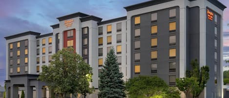 A multi-story hotel with a modern architectural design, featuring a prominent entrance with a glass door and a landscaped front yard with various trees.