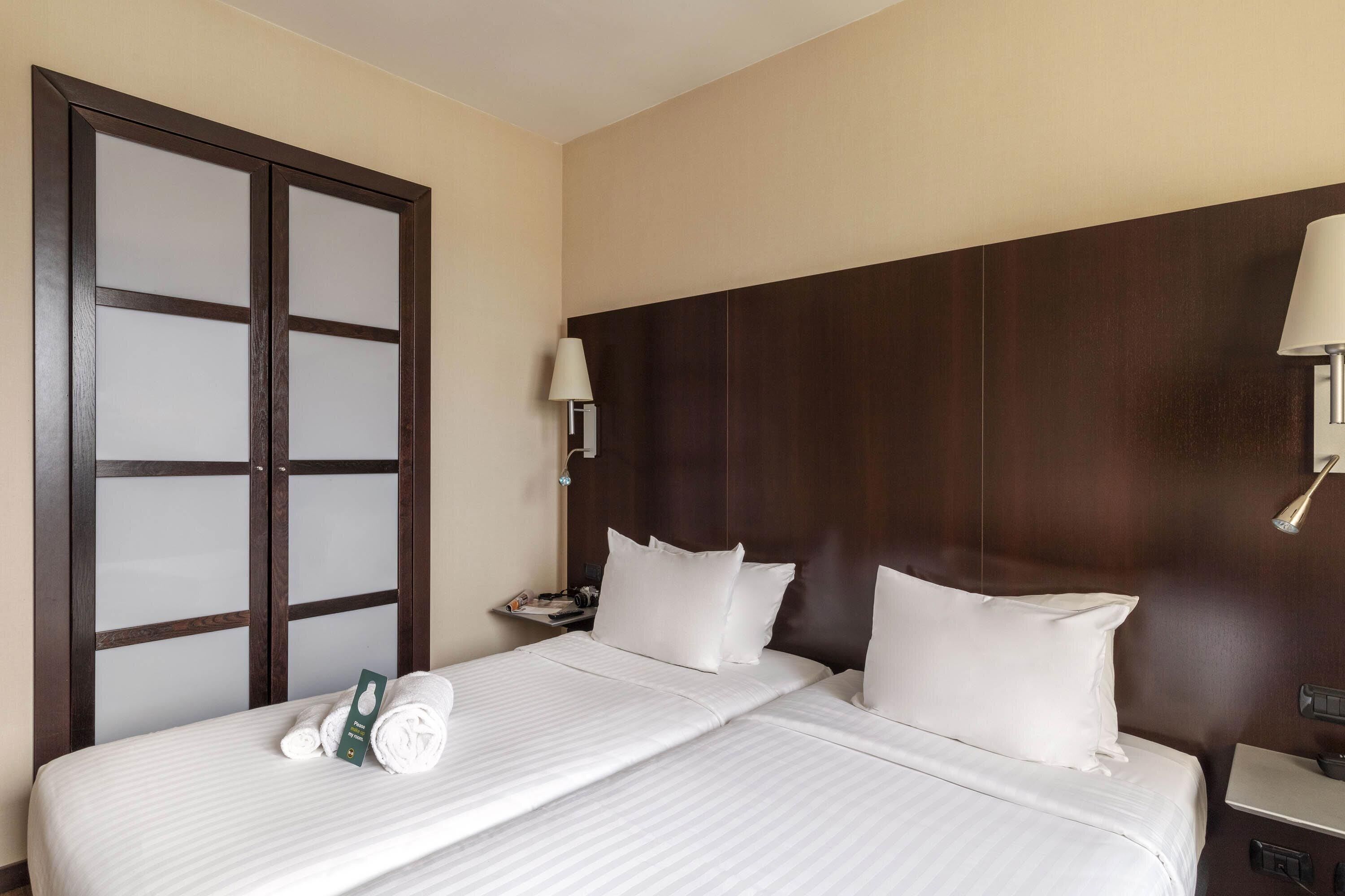 A hotel room with a large bed, two white pillows, a nightstand with a lamp, and a wooden headboard.
