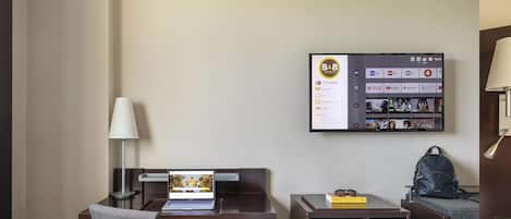 A hotel room with a desk, chair, laptop, lamp, and a TV mounted on the wall displaying a menu.